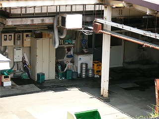 いくつかの一斗缶に突っ込まれたたくさんのいろんな傘、壁の電気メーター、古い縦長のロッカー、ビールのアルミ樽、その他。雑然とした駅事務室裏。