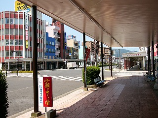 影を作っている平らなアーケードからグリーンベルトのある4車線道路を見る。道路の向こう側には4,5階建てのビルが並ぶ。
