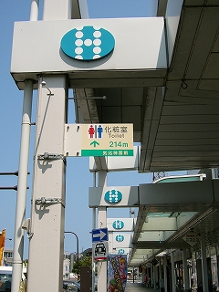 アーケードの柱の上のほうに飛び出すように取り付けられた横置きの長方形の鉄板製の看板。クリーム地に男女のトイレのマークと「化粧室　Toilet」214mと書かれ、その下の細長い緑地に「気比神宮前」と書かれている。