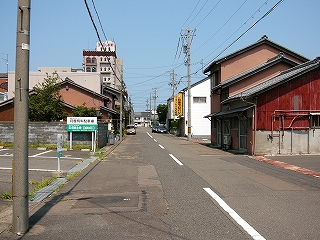 左の手前には駐車場があり、ほかは朴訥な住宅が並ぶ2車線道路。路面はつぎはぎだらけ。
