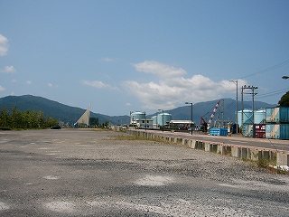 砂利の敷地。右手には60cmぐらいの段差の上に2車線の道路。右奥には大きなタンクやコンテナ、クレーン。