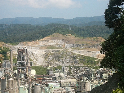 山肌がすっかり削られて白い地質が見えている。手前には複雑なパイプを張り巡らされた工場。