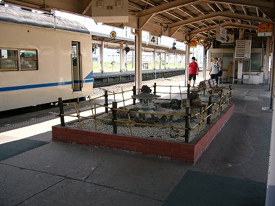 クリーム地に水色の線が窓の下に入った先頭車の乗務員室入口を赤レンガで低く囲われた枯山水と共に。列車の撮影者は二人いる。