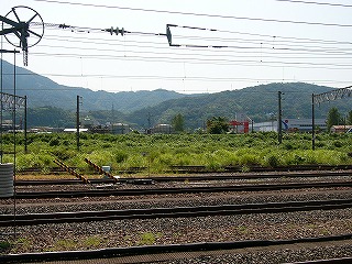 5本ぐらい向こうの線路から雑草が茂り、緑がもくもくと茂っている。その茂みの向こうに大きなガスタンクののようなものが2つ、その右に平たい工場、さらに向こうはまたもや山々。