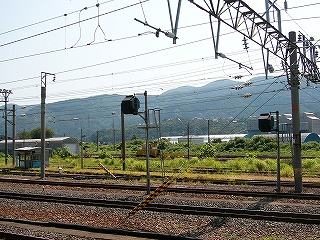 緑の雑草広がる留置線、その向こうにかまぼこのような建物が横長にあり、さらに向こうは緑の高台に何件か家があり、さらにその向こうは山々が横長に連なる。