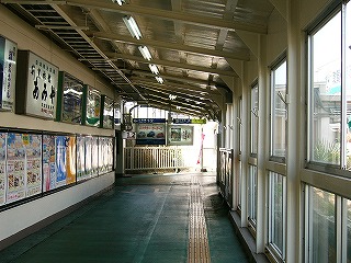 右に傾いた屋根。左は白壁にずらっと広告灯が並び、その下には紙のポスターがずらっと並ぶ。右は足元から天井までの窓ガラスだが、いったん真ん中ほどあたりでサッシは分断されている。床は緑色で、暗い黄色の点字ブロックが床の右寄りにまっすぐ伸びる