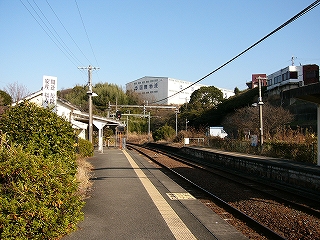 左手に成長したつつじの植え込み、右手に伸び行くホームと線路内。少し遠くの高い位置に木々に囲まれた運送会社の大きな建物。