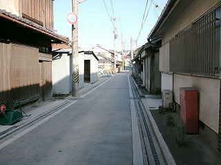 1.5車線の白っぽい路面の道。両脇には低い住宅が立ち並ぶ。