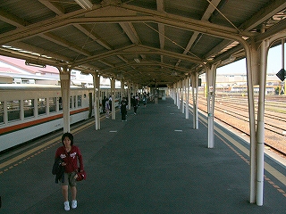 アスファルトのホーム。広い上屋。階段途中から見て。左の乗り場には列車が停車している。