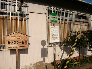 左にあいさつ一番と書かれた明るい木の立て札、右に緑色と白色でデザインされたバス停。