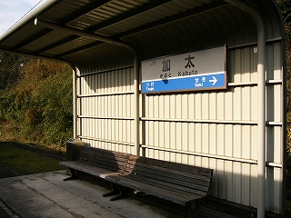 波打たせた金属の白い板でできた壁が屋根になるよう1ほぼ直角に曲げられて作られた待合所。その屋根の下には長椅子が二脚連られねられている。