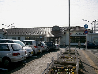 目の前には縦に並ぶパンジーの植えられた大きな正方形の木枠のプランター、その先にオレンジ色の瓦葺の横長の駅舎。