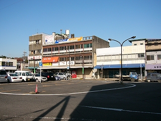 左手に先ほどの4階建ての旅館の建物、その隣に少し古い3階建ての横長の古い建物、その隣に2階建ての横に短い建物。建物の並びの前は駅前のロータリーだが、人は誰も歩いていない。