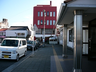 ステンレスの長い二本の四角柱で支えられた駅舎入口ポーチを真横から見て。奥の方に4階建ての赤黒いビル。