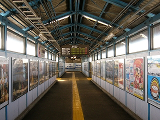 濃い灰色のコンクリートボードを濃い水色の鉄骨にはめ込んでできた跨線橋内。両脇にはポスターがずらりと並ぶ。