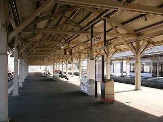 背の高い山型の上屋が覆いかぶさる幅の広いホームを上屋の下から見渡したようす。真中付近に椅子と時刻表と天井から吊られた駅名表があるだけ。