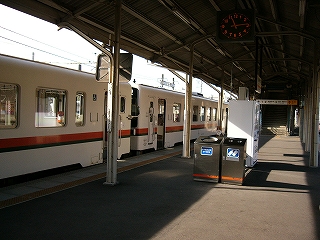 ホームの上屋の下から白地にオレンジと緑を重ねた細いラインの入った二両編成の列車を途中から見て。ホームの真中にはゴミ箱と自動販売機が置かれている。