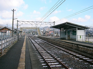 遠くまで伸びる線路内とホーム。遠くに跨線橋とトラス架線柱が見えている。線路内を挟んだ右手にはホームにある大きな屋根を持つ開放式の待合所が近くに見えている。