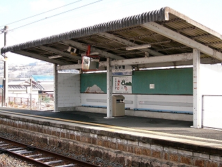 線路外に大きく傾いた、面積の広い波板の屋根をもつ開放式の待合所。内部の壁には緑色の横長の掲示板が設置されている。壁は白色、屋根はカビた灰色。