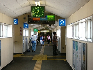 白い天井と壁の跨線橋内。天井は切妻になっていて、床面はアスファルト。通路の幅は横に並んで4人が通れるぐらい。階段下り口付近には青地に白い字で数字が書かれた電照式番線番号や4色に色分けされたフラップ式番線案内が天井付近に設置されている。