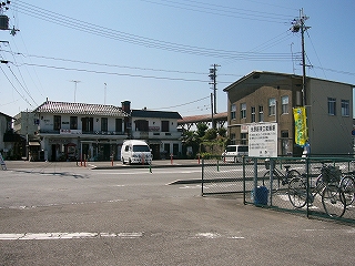 人も車のとおりもない、横切る道路を挟んで、左手に二階建ての横長の建物、その建物の右手に、少し手前ずれて立っている焦げ茶の壁の2階建ての建物。