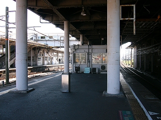 手前にホームの両脇に立つ白くて太い二本の柱。それが上にあるコンコースを支えている。コンコースは左の線路内には突き出ておらず、立ち位置はそれほど暗くない。二本の柱の奥には白い簡単な詰所が置かれている。