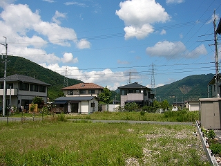 手前に雑草の生えた空き地、その奥に3件の家。ずっと奥に夏山と青空。