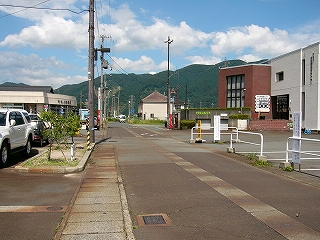 赤茶けた道。少し遠くに真新しい建物の郵便局、右手に赤いレンガ壁と白い壁で造られたセンター。