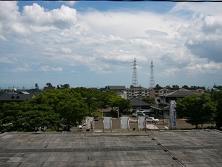 すぐ下に駅舎の灰色と黒に薄汚れた陸屋根、その先に緑の並木と少しの住宅が見える。