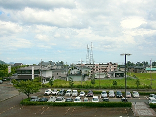 路面が赤茶けた駐車場。その奥には緑の空き地があり、その奥から住宅が始まっている。