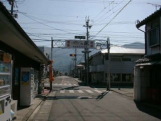 信号のある小さな十字路。交差点に出る直前に簡易なゲートがある。脇に自販機のある民家。
