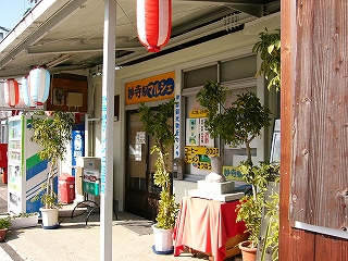 いくつかのちょうちん、黄色地に青い字の看板、観葉植物の並べられた店の前。