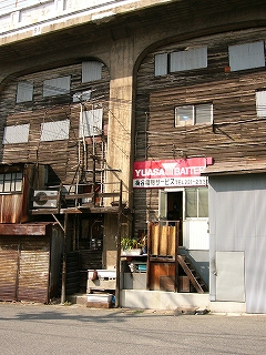 高架下に目いっぱいの高さで作られたこげ茶色の木造の倉庫のようなもの。多くの窓は灰色の板で塞がれている。