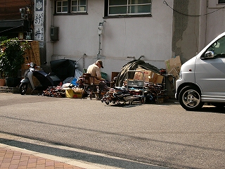 路上脇にたくさん集められた傘に囲まれて、椅子に座りながら傘をまとめているおじいさん。