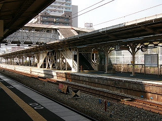 屋根のある隣のホームを右斜めに長く見て。少し遠くで跨線橋がそのホームの屋根の上で直角に交わり、交わったところで屋根と壁のある階段がホームの屋根を突き抜けて、ホーム上に口を開いている。