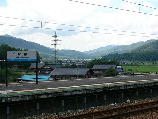 隣のホームの地上に立った駅名標とその向こうに見下ろせる田、そしてずっと向こうに家々が広がり、周りは低山に囲まれる。