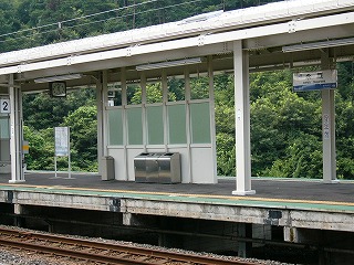 白い鉄骨に支えられた上屋、ホームの中央辺りにあるガラスの入った風よけ、右に上屋からぶら下がる駅名標、背景は木々の覆う山裾。