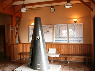 上の写真の右側。背の高い円錐の彫刻の後ろに明るい木の色の長椅子、その上に時刻表。