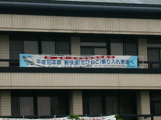 横断幕。上に細く赤地に白字の文字、その下に大きく水色の字でうたい文句。