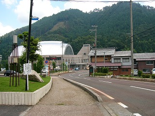 向こうにカーブした道の左脇に体育館のような蒲鉾屋根を持っている新しい建物。向こうは山。