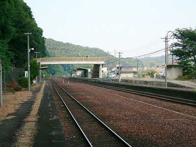 左に暗い緑の山肌と日陰になったホーム、そして広い停車場、屋根のない跨線橋、さらに下り線ホーム。