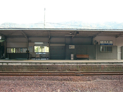 上り線ホームと駅舎を向かいのホームから見て。駅舎から伸び出てきた横に長い屋根がホームを暗くしている。駅舎のホーム側の壁は鼠色でこれがなお一層暗くしている。改札口は丸く太い柵とステンレスの大きなきっぷ箱で構成され、改札口の上には蛍光灯が一本ともっている。
