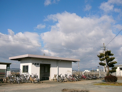 床面積より広い陸屋根をつけた簡易な駅舎を斜めに見て。少し離れた所に成長した背の高い植木が一本植わっている。