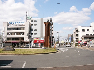 中央部の緑の芝生のスペースに背の高い木製のモニュメントが建っている。その向こうにKECゼミナールのビル。