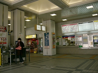 奥に有人の出札口と券売機が並んでいる。手前に公衆電話で電話する女性がいる。