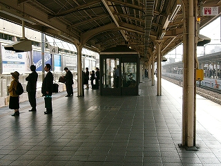 濃いねずみ色のタイルを敷き詰められた広いホームの先に新しい待合室のある風景。