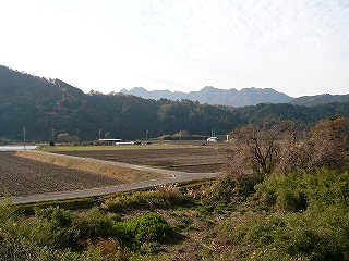 手前は枯れ草や枯れ木で荒れた敷地、その奥には休んだ田んぼが見えるが、少し遠くに山が迫ってきている。