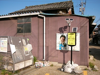 左手に背の低いステンレス製の扉つきのゴミを格納するゴミ入れ。右手に一本の棒に支えられた黄色い小さな縦長の看板があり、その上にソーラーパネルが取り付けられ、その看板からさらに棒が延びたその上にはカメラが取り付けられたものがある。