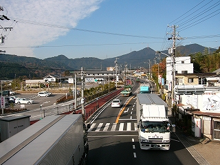 歩道橋から鈴鹿峠方面に見た国道1号。道は遠くで少しゆるくカーブし、遠くには少し高い山並みが横たわっている。眼下左手には駅前の駐車場とその奥の道の駅が見える。