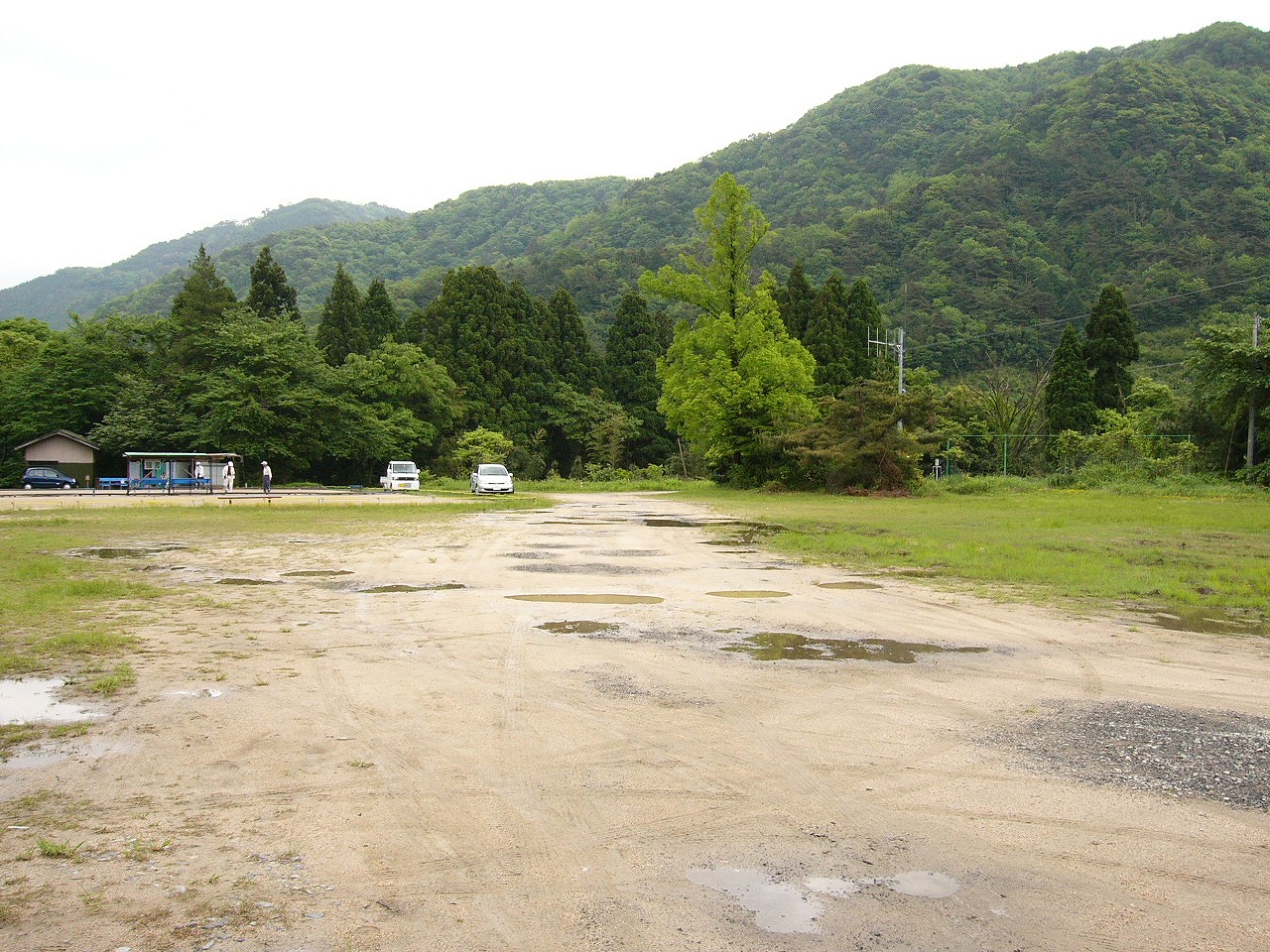 水たまりがいくつかあるグラウンド。真ん中は車3台分ぐらいは草が生えていないが、残りの両脇は草が短く生えている。奥に山。
